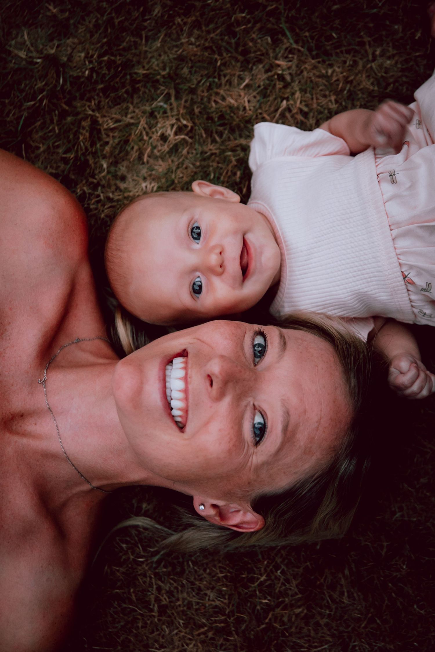 Overhead view of a baby and caregiver lying on grass sharing a joyful moment.