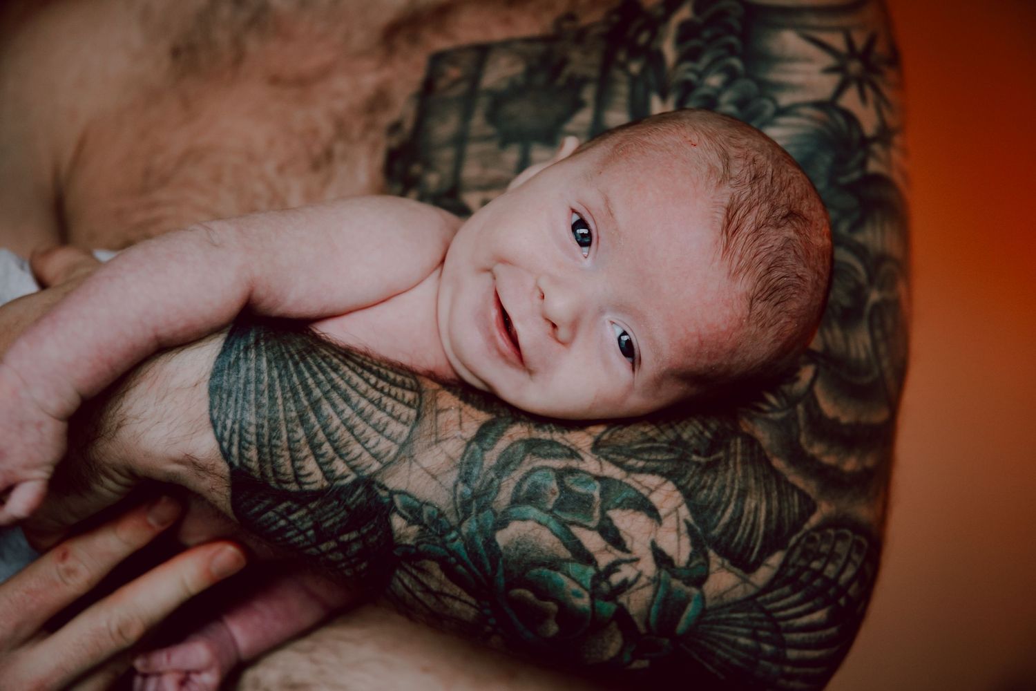Close-up of an infant being cradled in tattooed arms against an orange background.