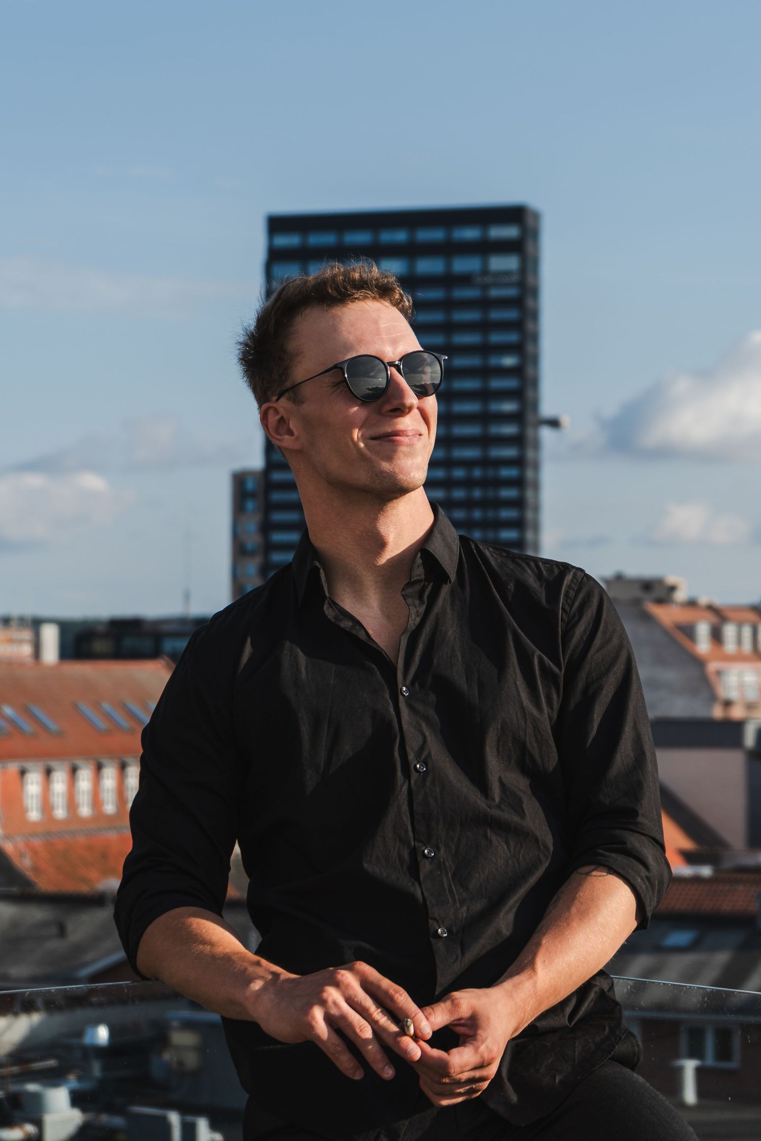Smiling young man in black shirt and sunglasses posing on rooftop with city skyline on sunny day.