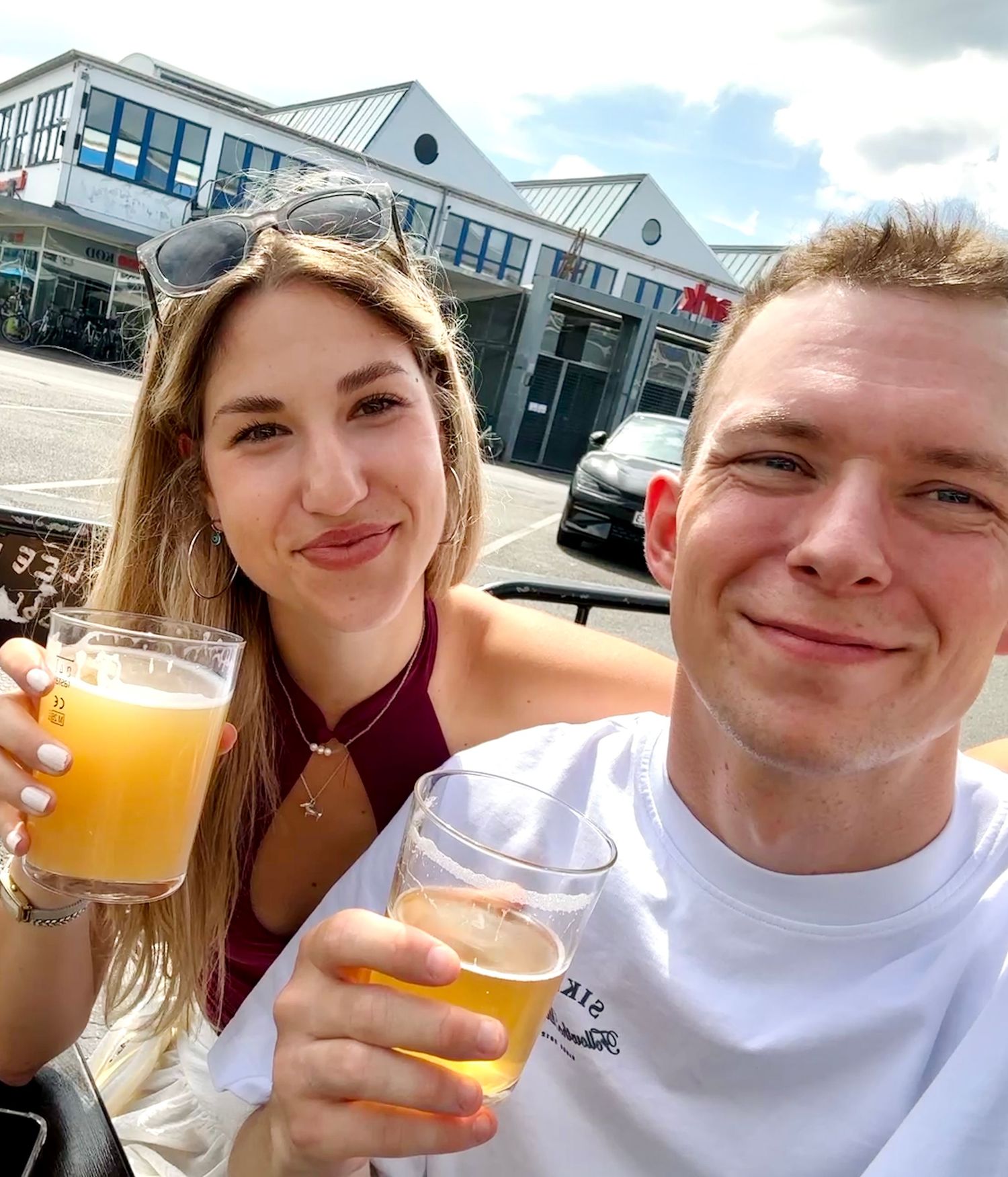A smiling couple enjoying drinks outdoors on a sunny day, woman holding orange juice and man holding beer.