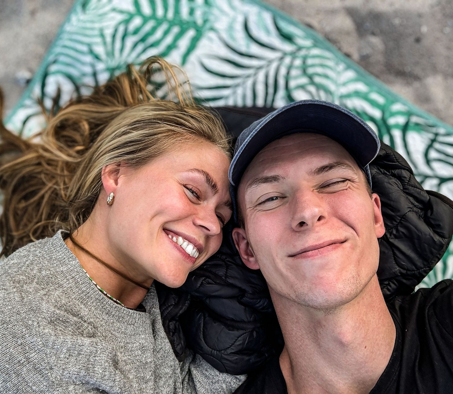 A close-up selfie of a couple lying down together and smiling with palm print decorative pillows in the background.
