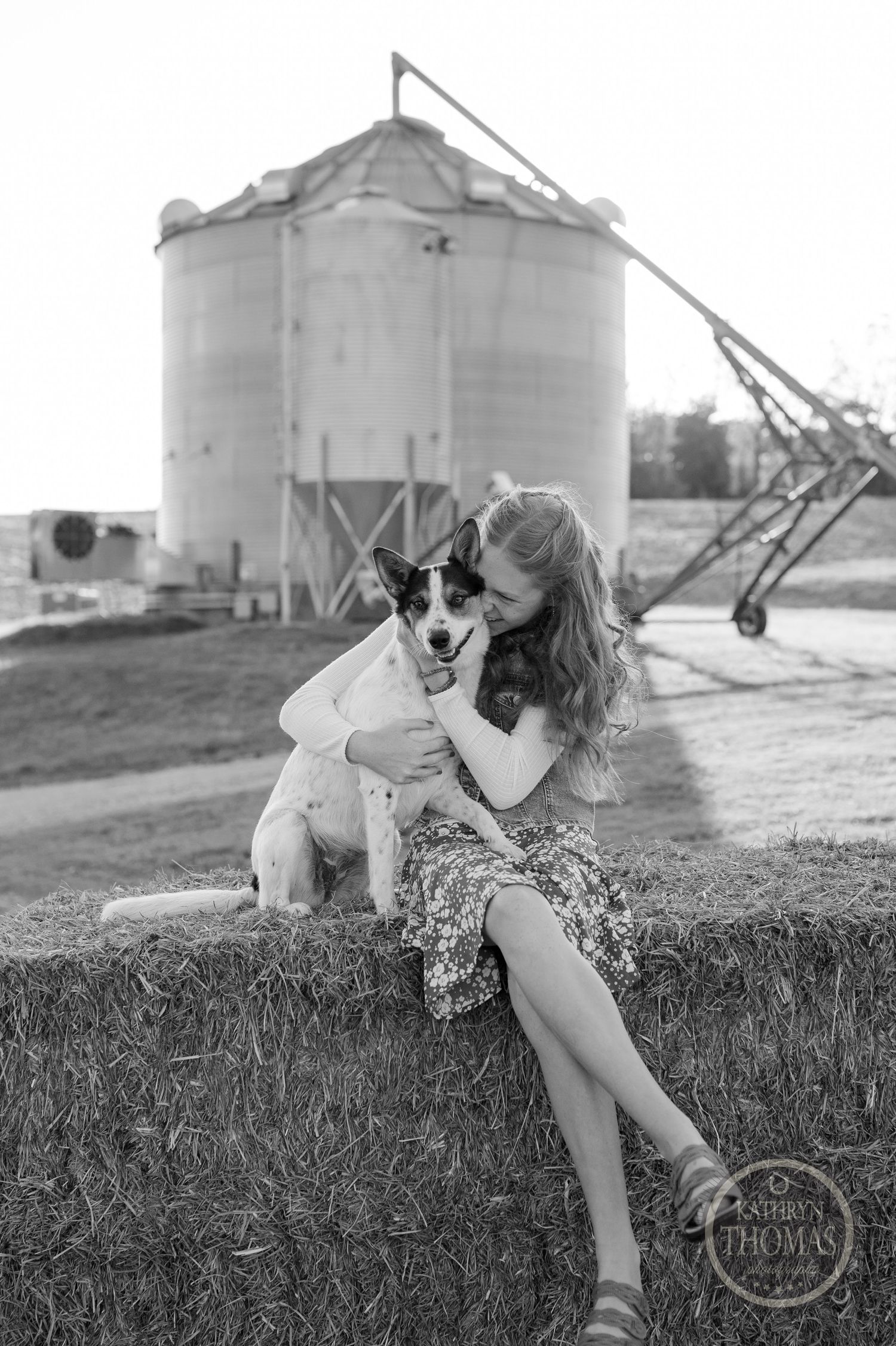 Black and white photo of a person and dog sitting on hay bale with grain silo in background at sunset.