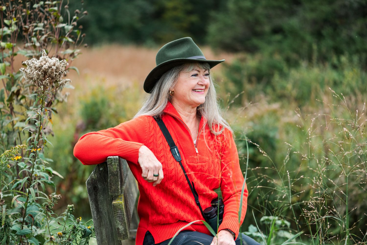 Someone wearing a bright red sweater and green hat laughs while sitting outdoors among wildflowers and greenery.
