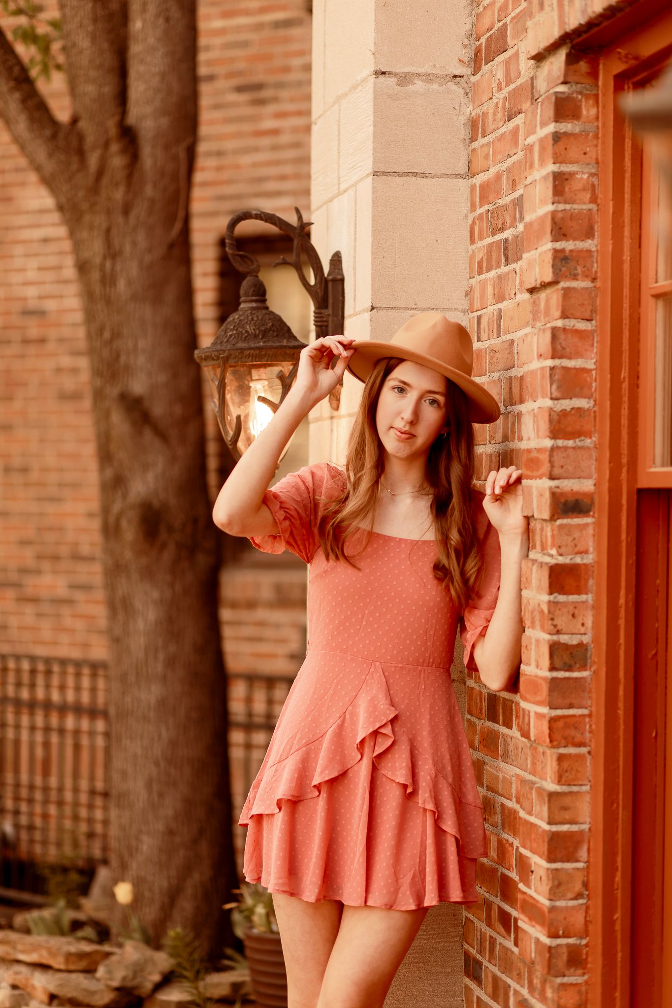 Romantic peach-colored ruffle dress and brown wide-brim hat outfit against rustic brick wall with vintage lantern.