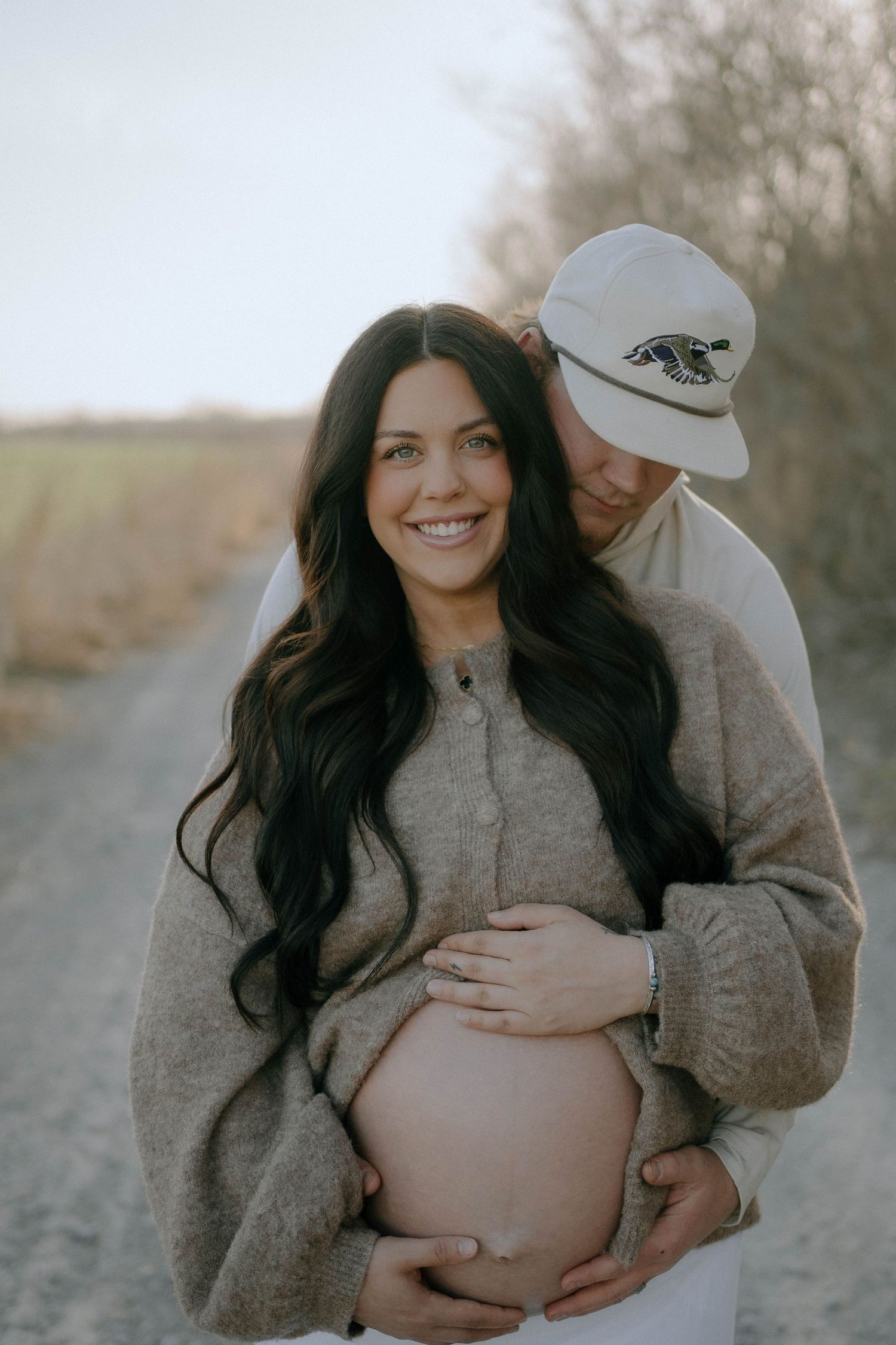 A heartwarming maternity photo of a couple in neutral sweaters embracing on a dirt path during golden hour.
