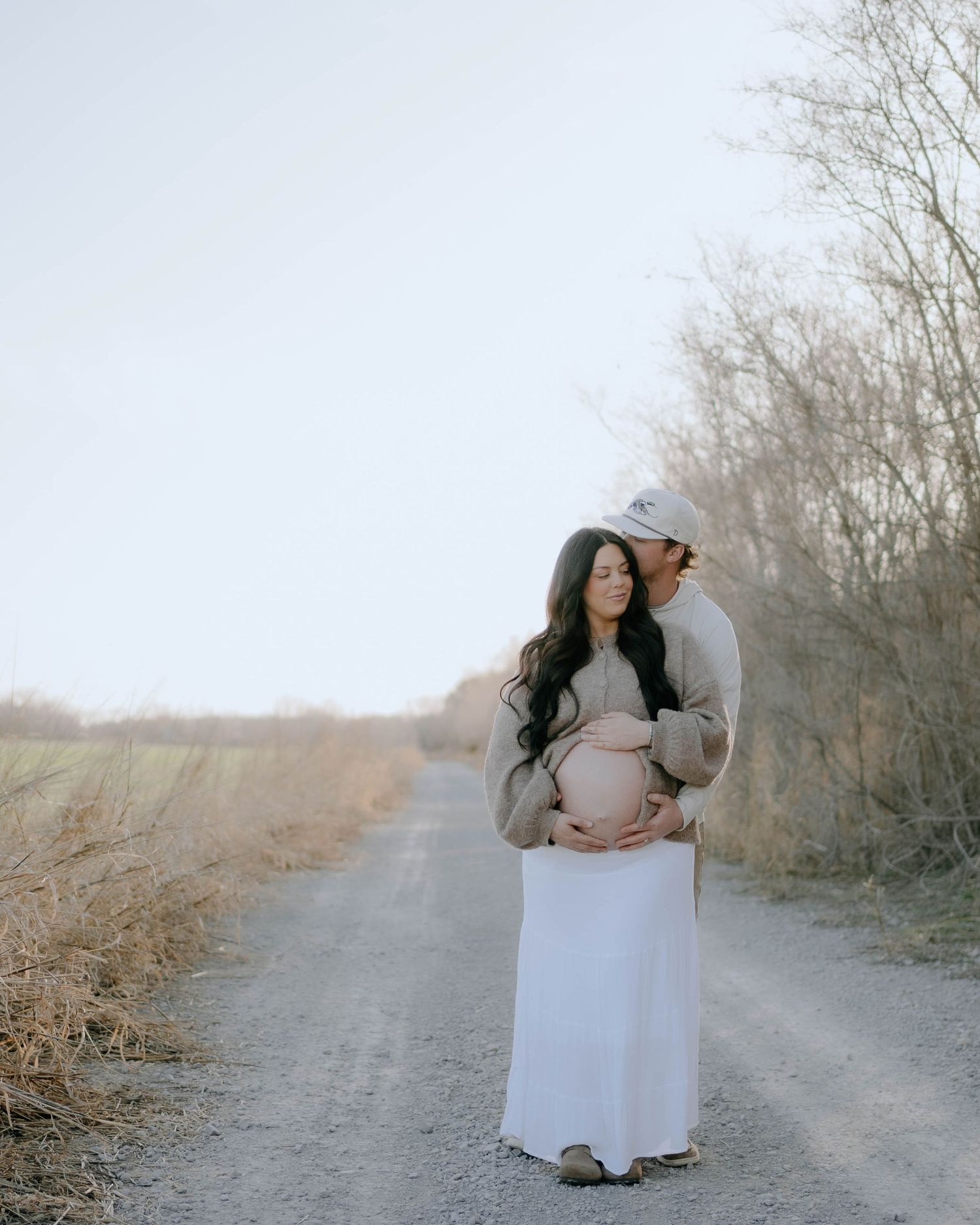 A couple stands together on a dirt path during a maternity photo session amidst bare trees on a misty day.