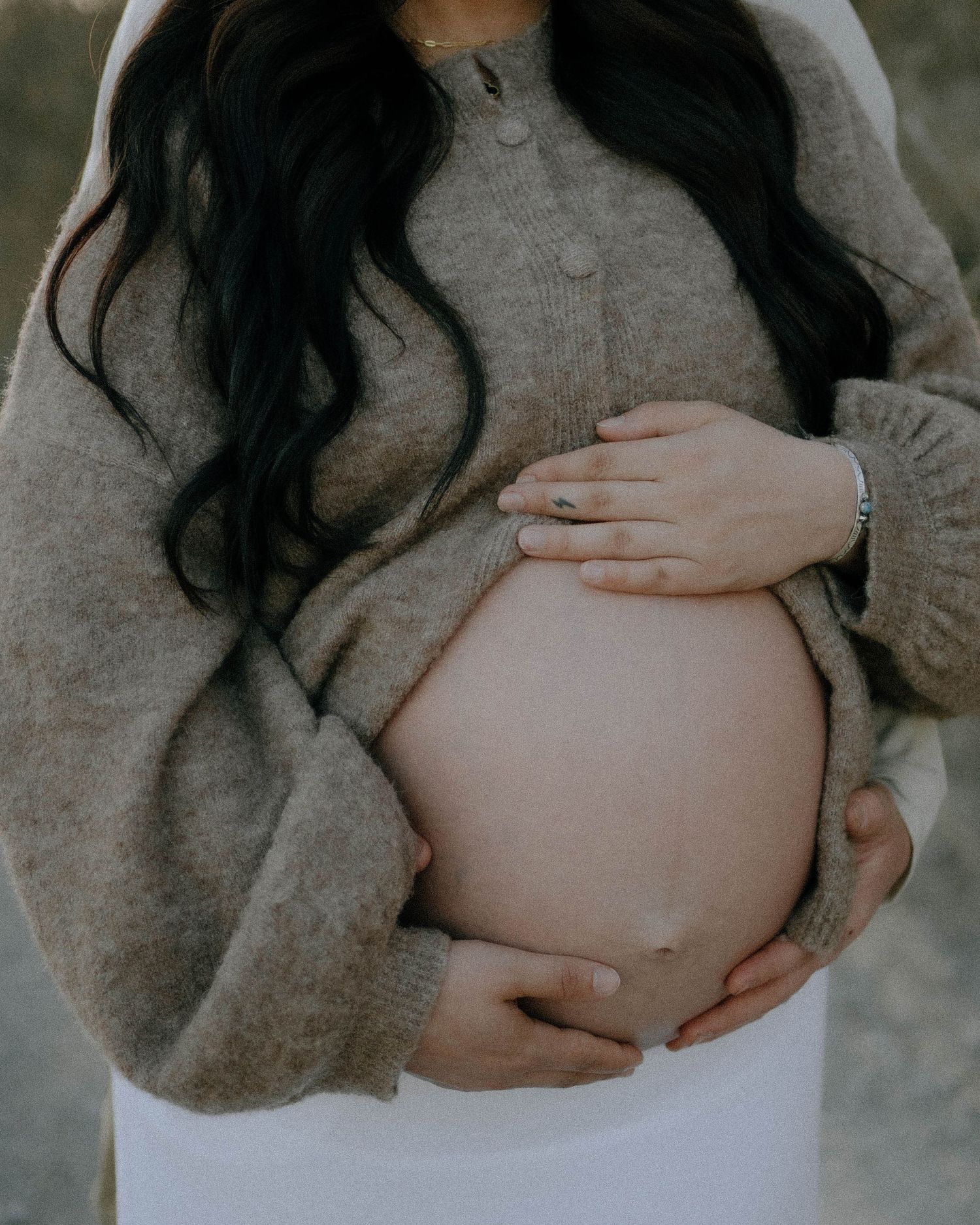 Expecting mother holds her bare pregnant belly while wearing a cozy gray sweater and white pants in soft light.