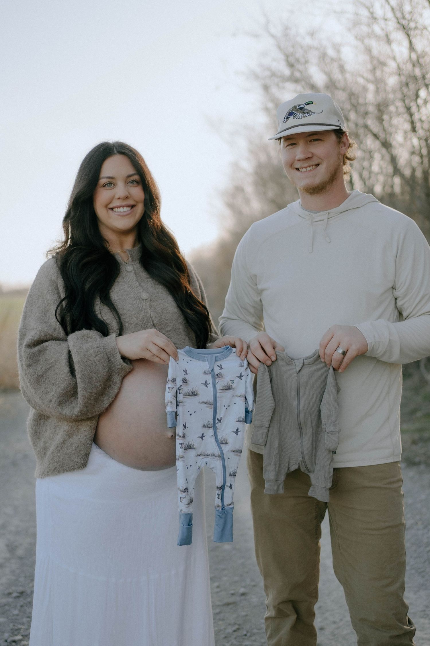 A couple outdoors holding baby clothes during a pregnancy photo shoot on a natural path in muted tones.