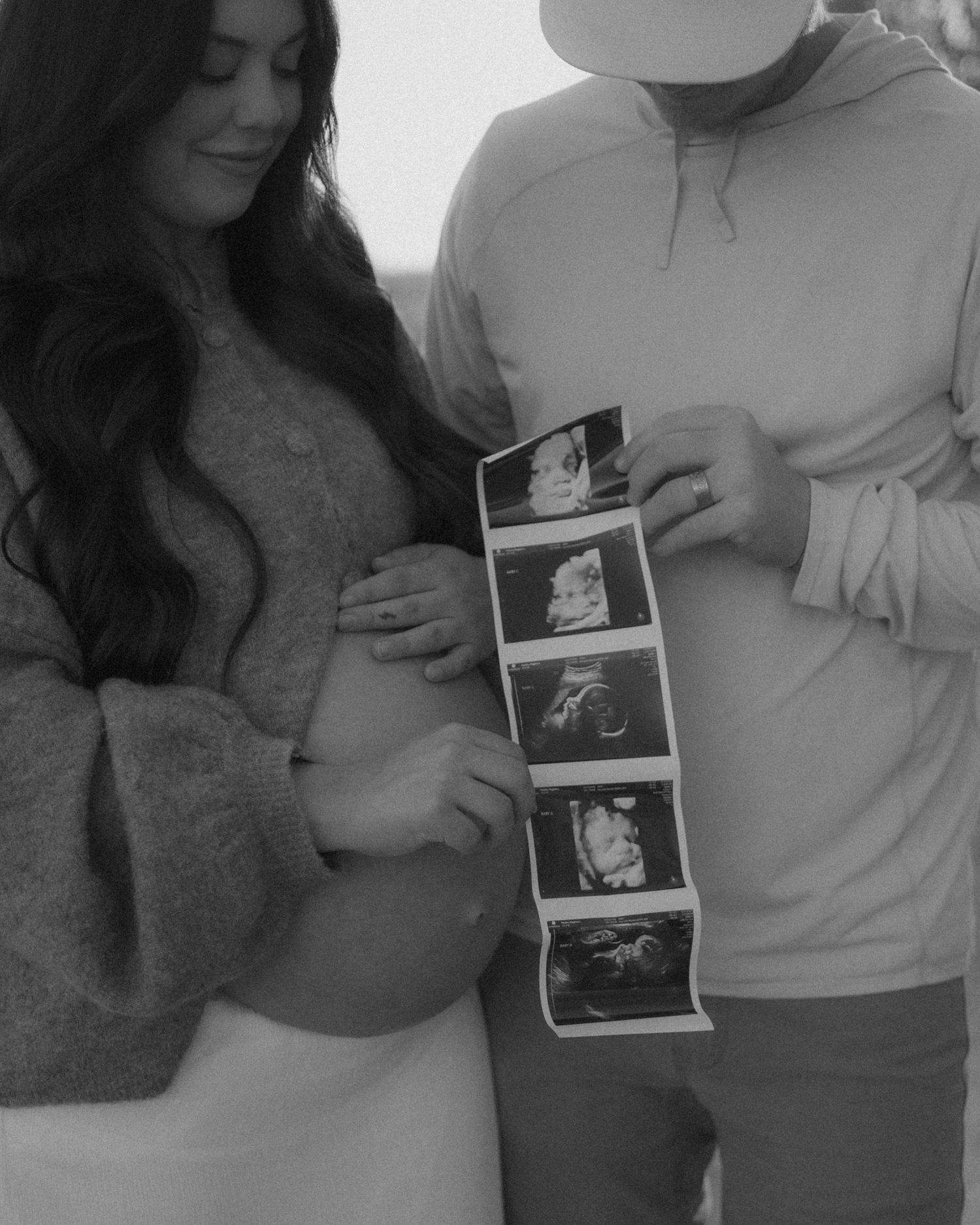 A couple holds ultrasound sonogram photos while the woman touches her belly in a black and white photograph.