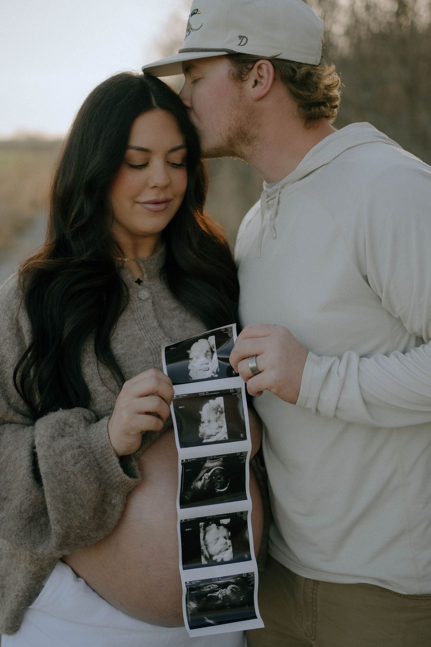 Couple sharing intimate moment while holding ultrasound photos of their expected baby during pregnancy announcement photoshoot.