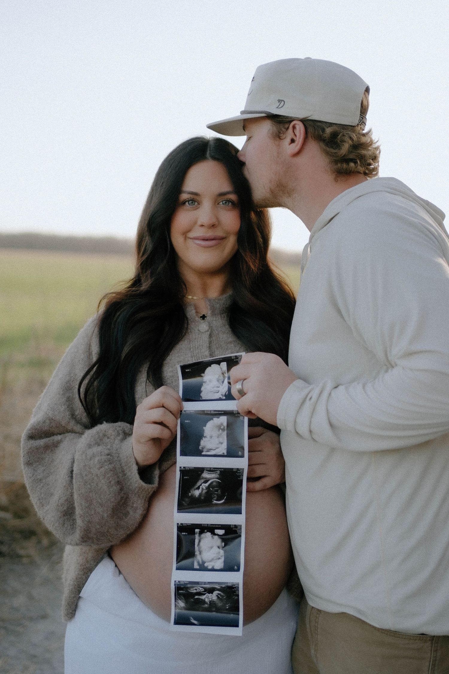A couple shares an intimate moment outdoors while holding ultrasound photos in a pregnancy announcement photo shoot.