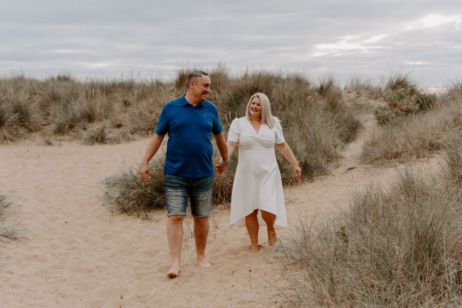 A couple are walking hand in hand on a beach. The man is looking over at his wife and smiling.