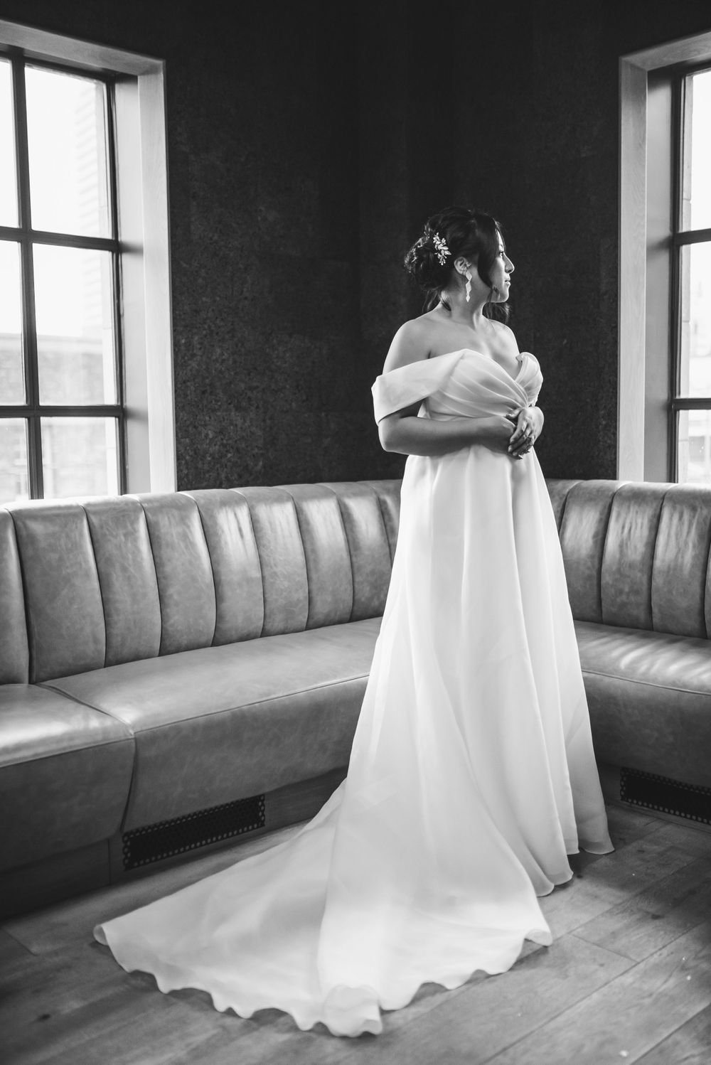 A person in an elegant off-shoulder white wedding gown stands between two windows next to a gray leather banquette.