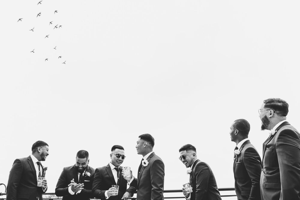 A black and white photo of groomsmen gathered together outdoors with birds flying in the sky.