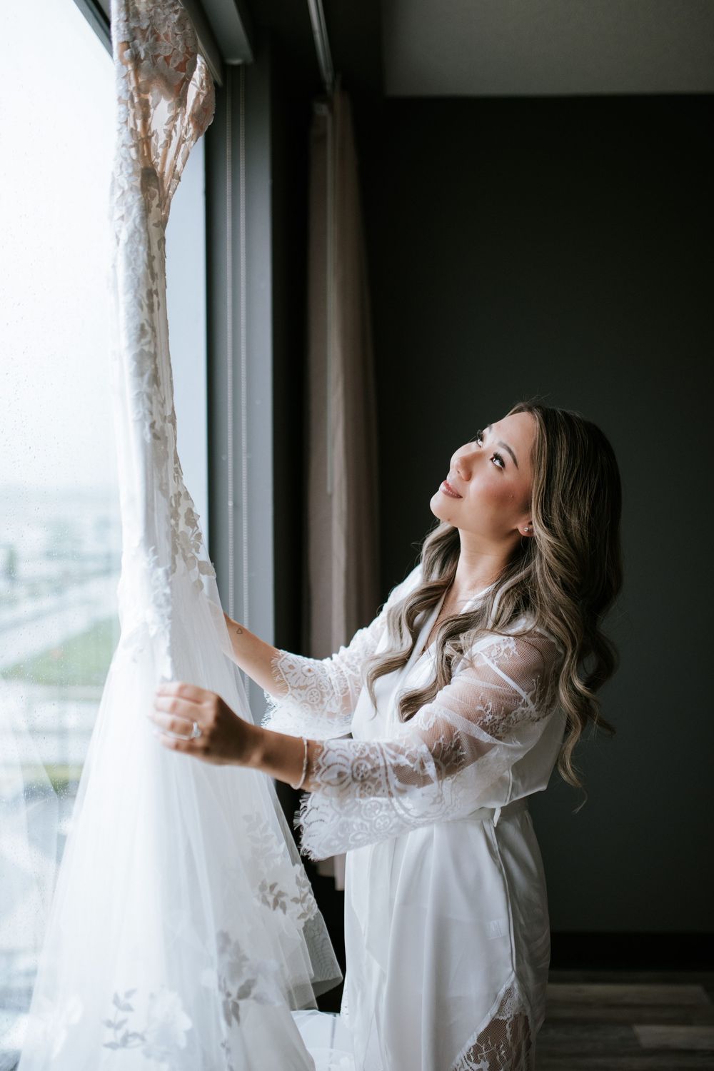 A person in a white silk robe admires a lace wedding dress hanging by a window with natural light streaming in.