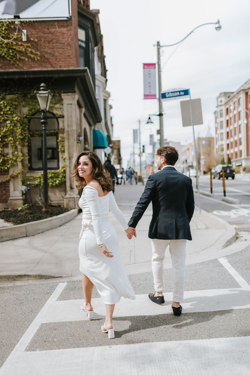 Stylishly dressed couple walks across urban crosswalk holding hands near brick building with vintage lamp post.