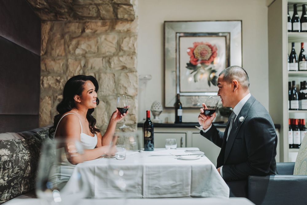 Two people share wine at an elegant restaurant table with stone walls and a floral print in the background.