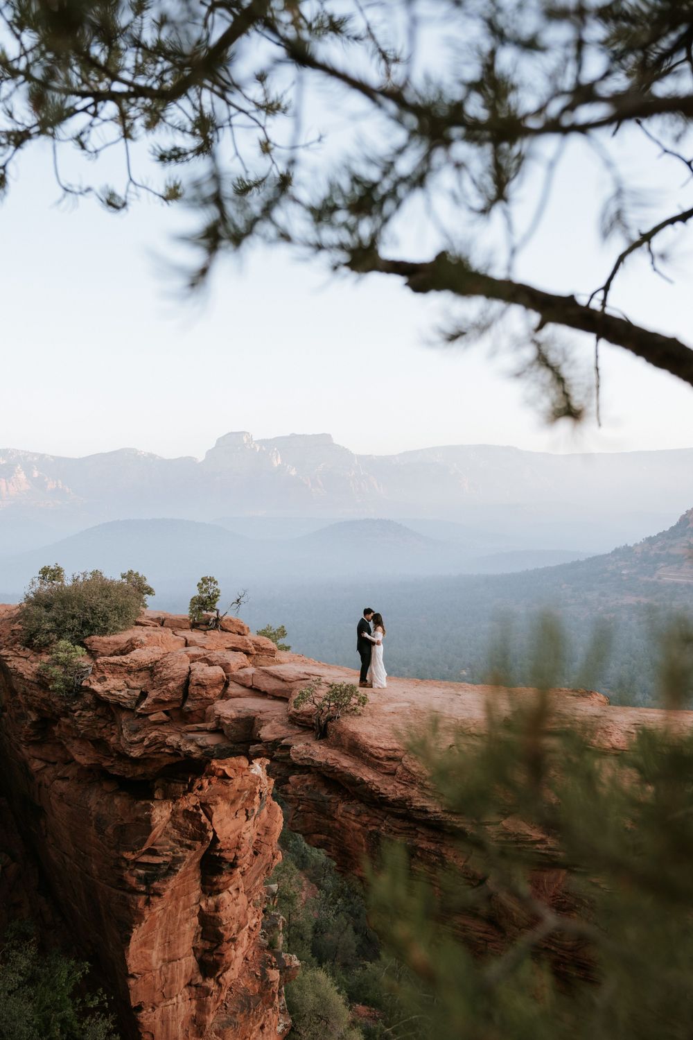 A silhouette stands on a red rock cliff edge overlooking misty mountain ranges in the distance through pine branches.