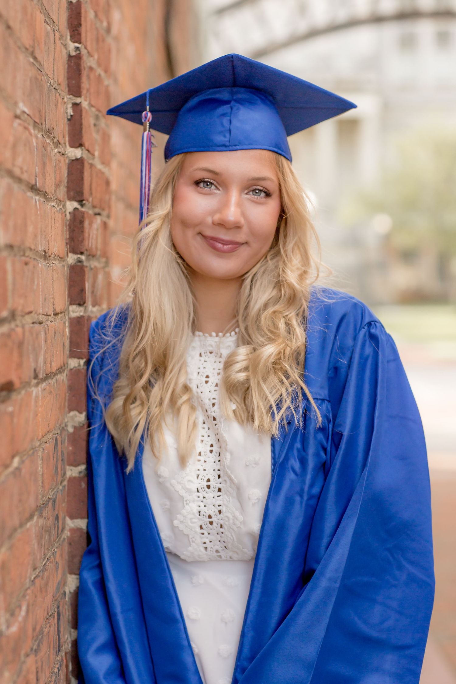 Smiling blonde woman in blue graduation cap and gown leaning against a brick wall.