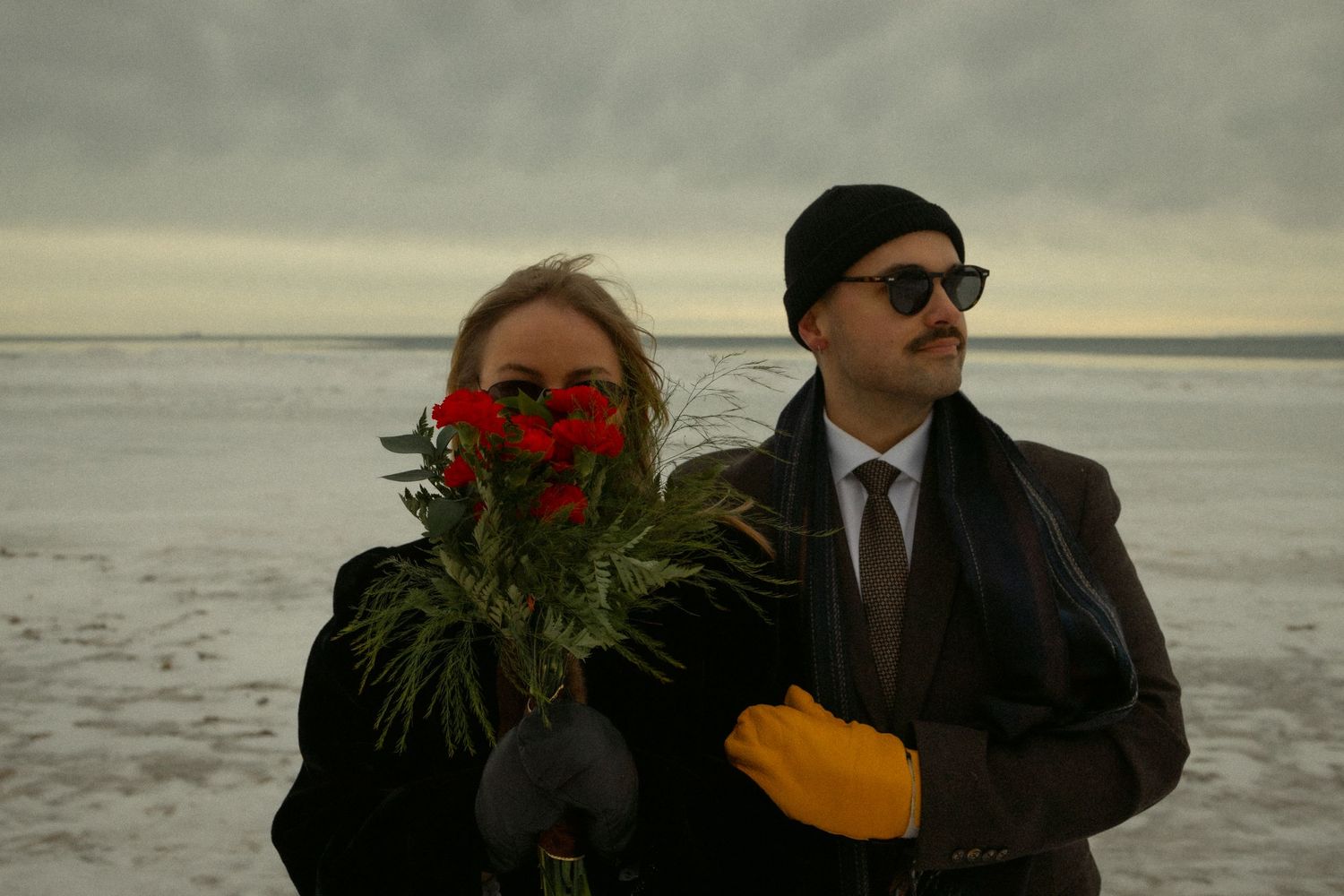 Two people stand on a beach at dusk, one holding red roses while the other wears sunglasses and carries a yellow folder.