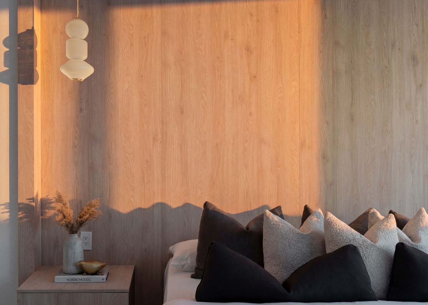 Cozy interior corner with sunlit wall, gray and black throw pillows on seating, and small vase with dried flowers.