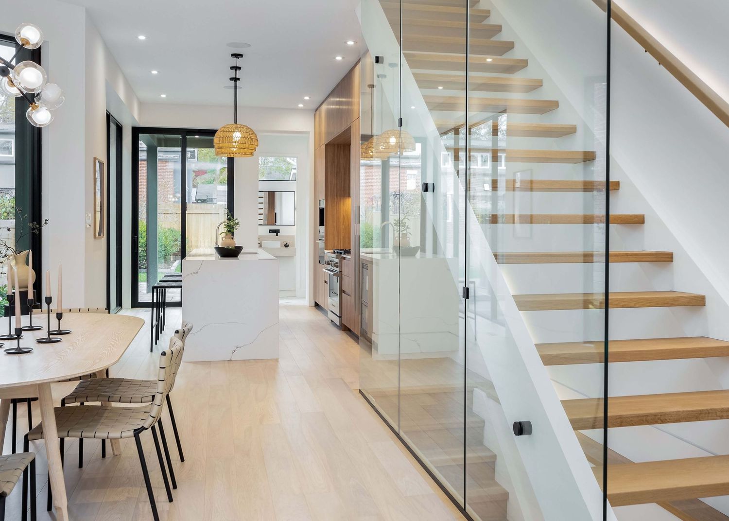 Modern home interior featuring floating wooden stairs with glass railing and light hardwood floors in an open concept space.