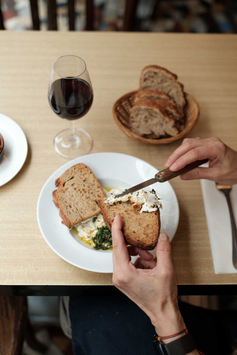 Hands spreading butter on rustic bread with wine and place settings on a wooden table.