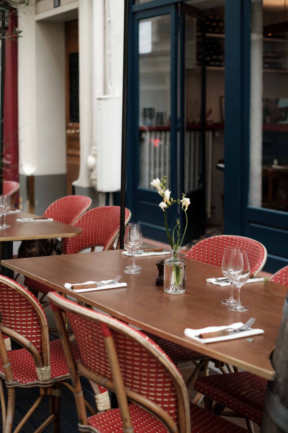 Outdoor cafe seating with red woven chairs and wooden tables set with wine glasses and small flower vases.