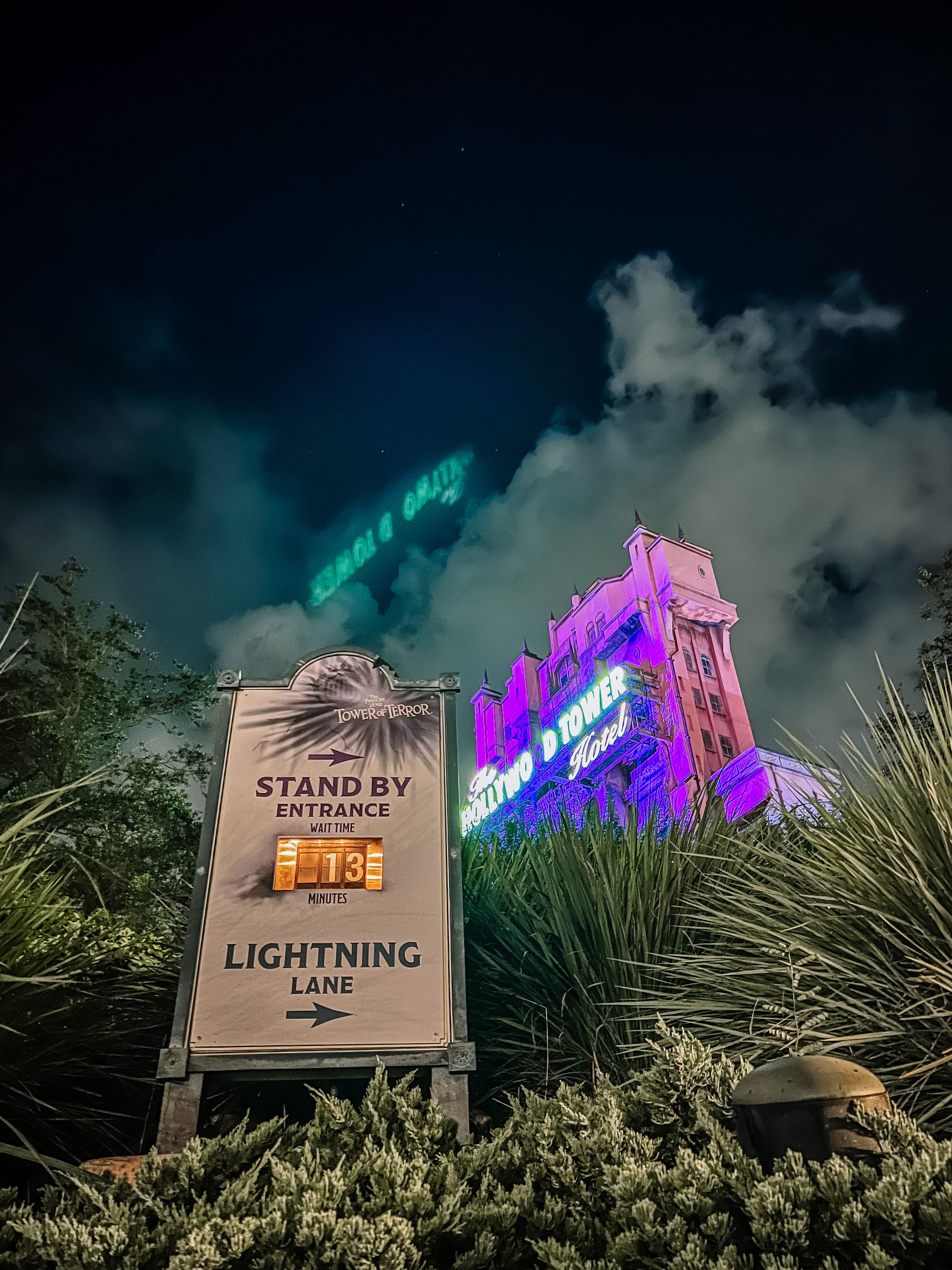 Theme park directional signs for Lightning Lane and Stand By entrances with colorful lighting effects at night.