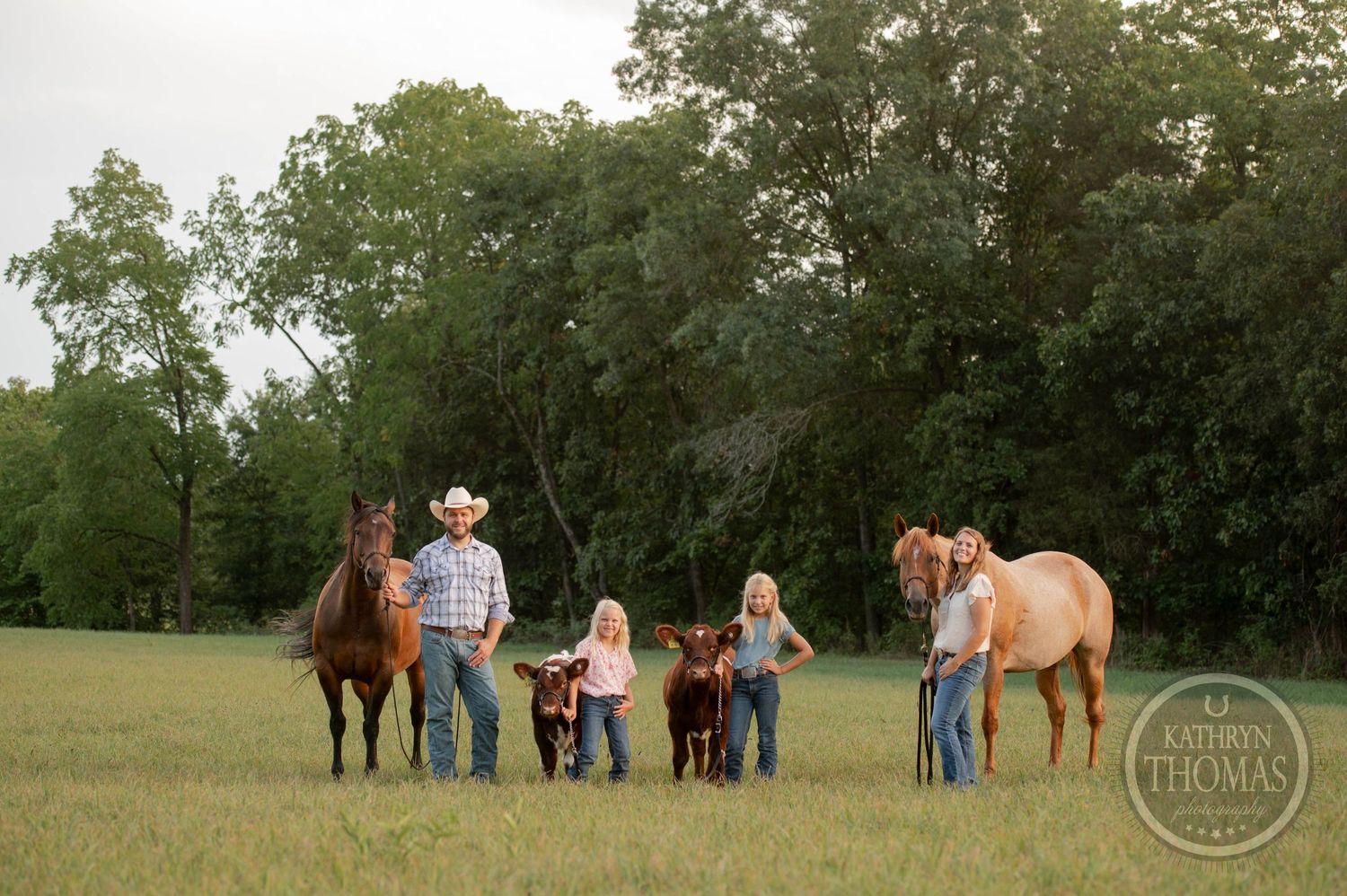 A family group walks through a pastoral field with horses and calves at dusk.