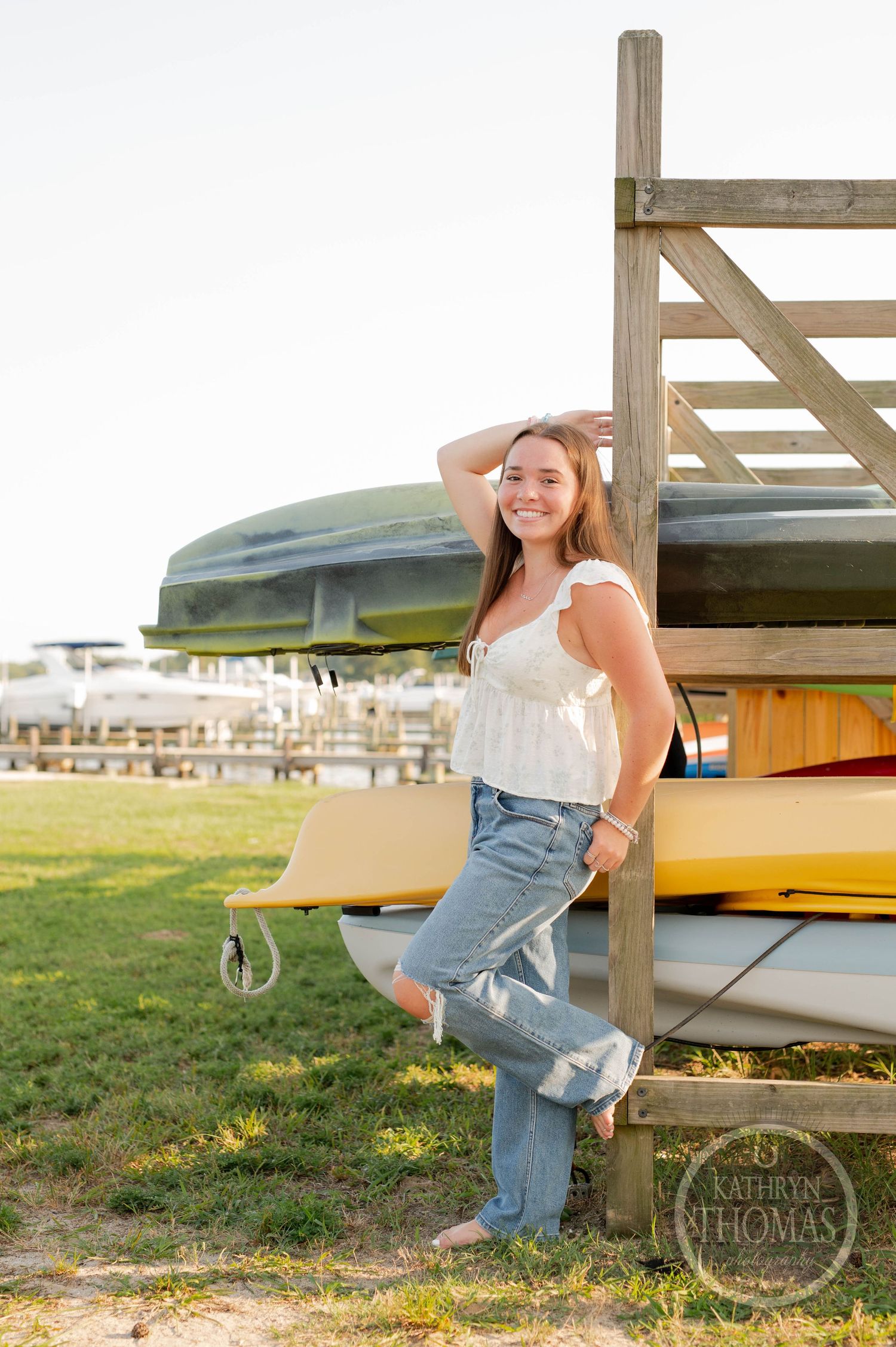 Portrait of someone in a white top and jeans posing by stacked kayaks on a wooden rack near the beach.