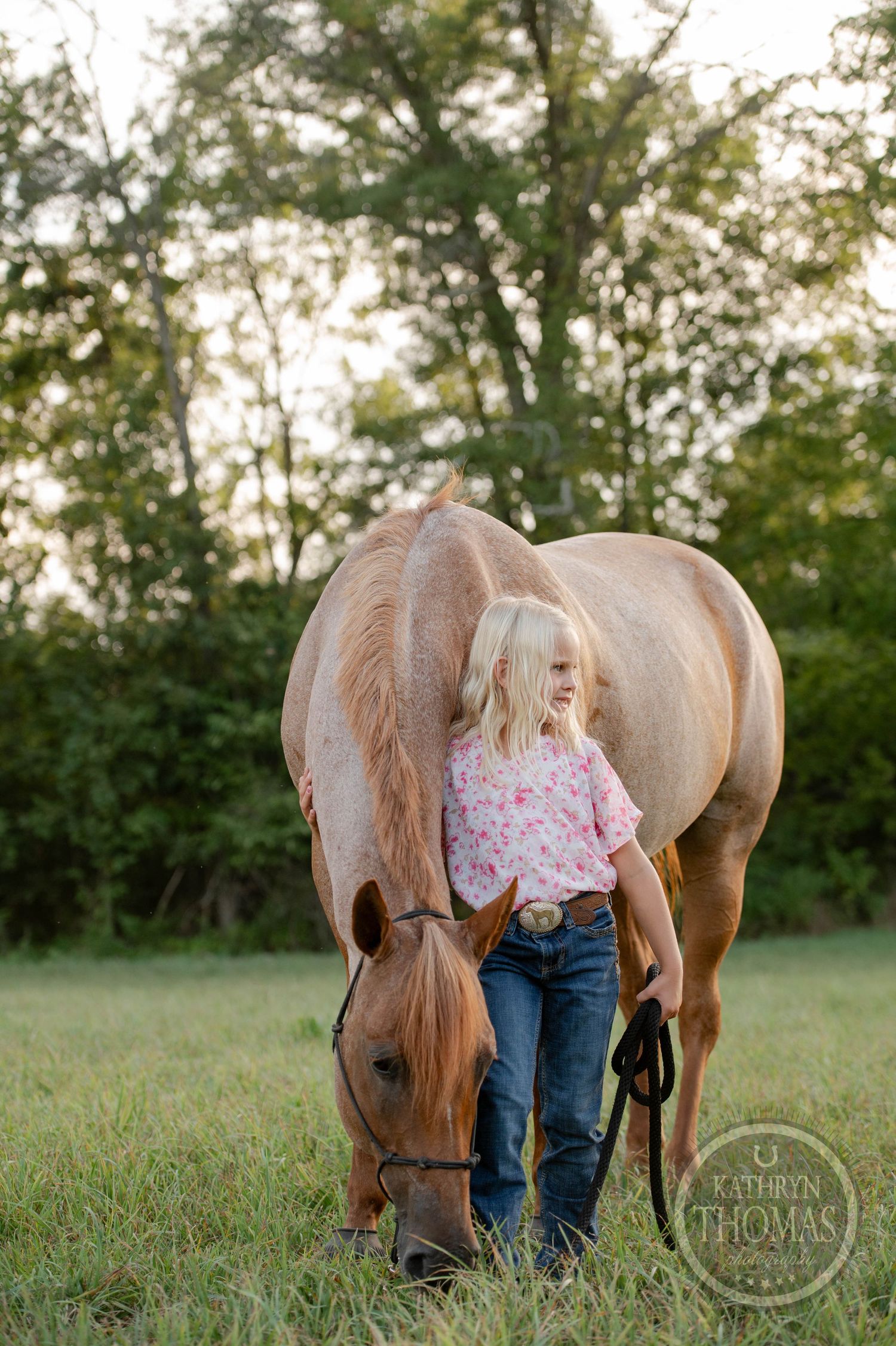 A person in jeans stands beside a grazing brown horse in a peaceful meadow setting.