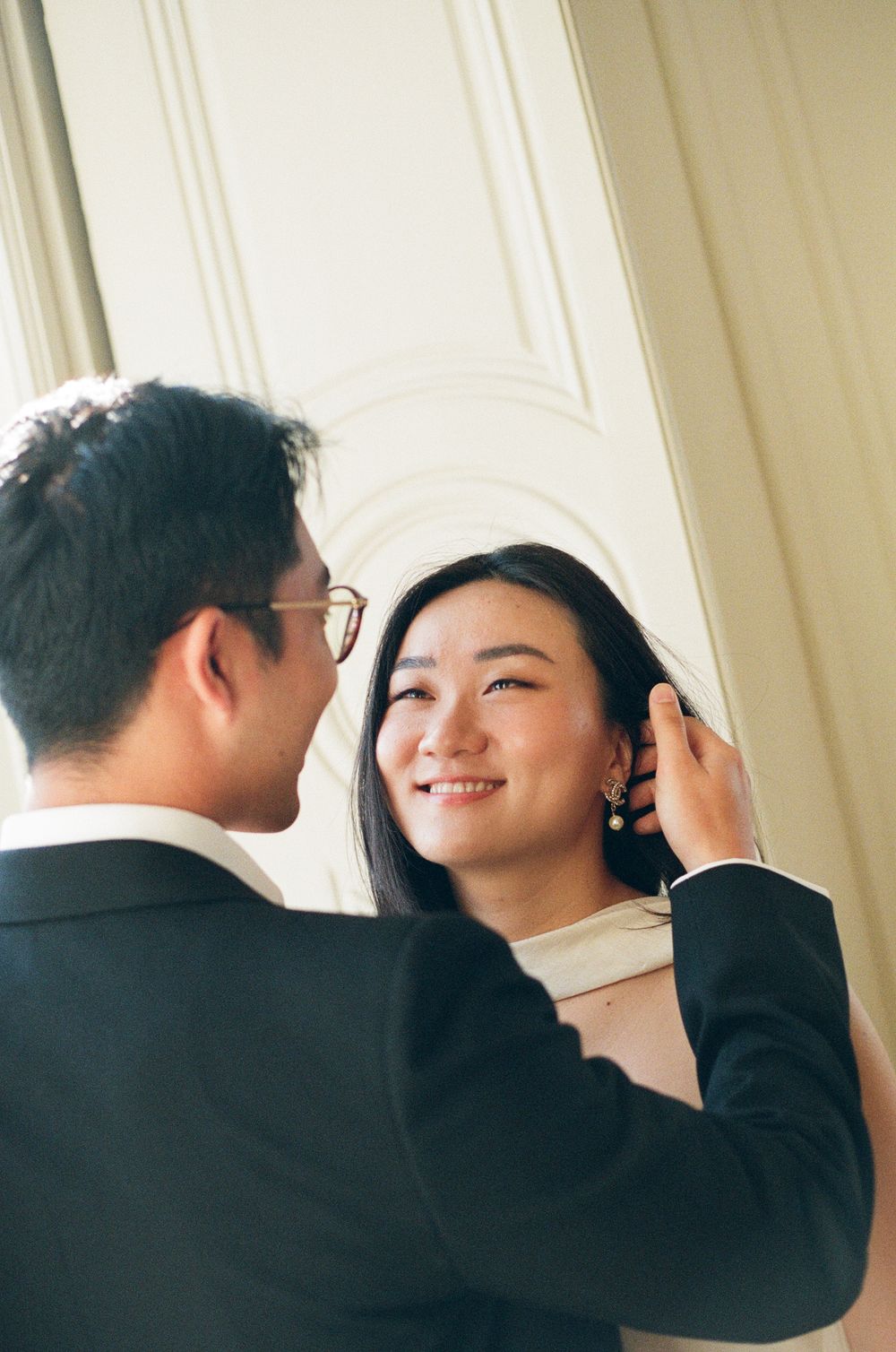 A groom in a black suit gently tucks hair behind a smiling bride's ear in an elegant white interior. paris pre wedding photoshoot film photo