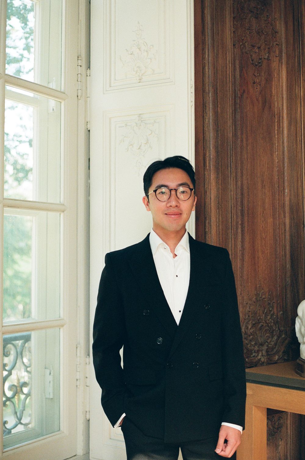 Young man in black double-breasted blazer and glasses standing by ornate white door in elegant interior. musee rodin film photos