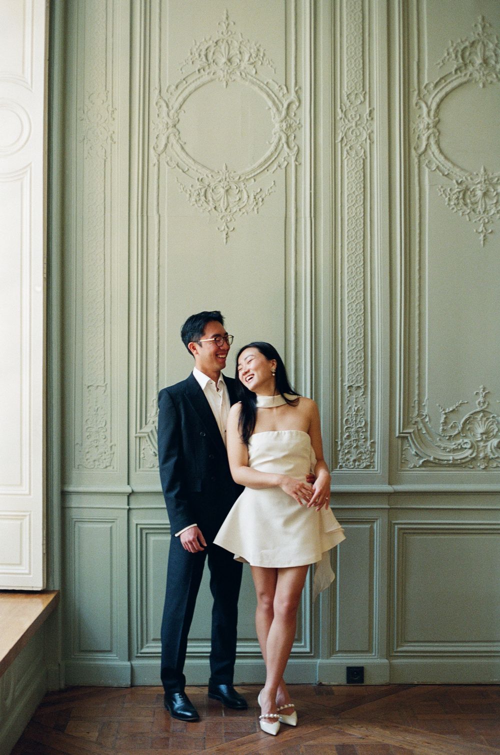 Couple in elegant attire poses before ornate sage green French molding wallt. paris film wedding photographer, editorial photography