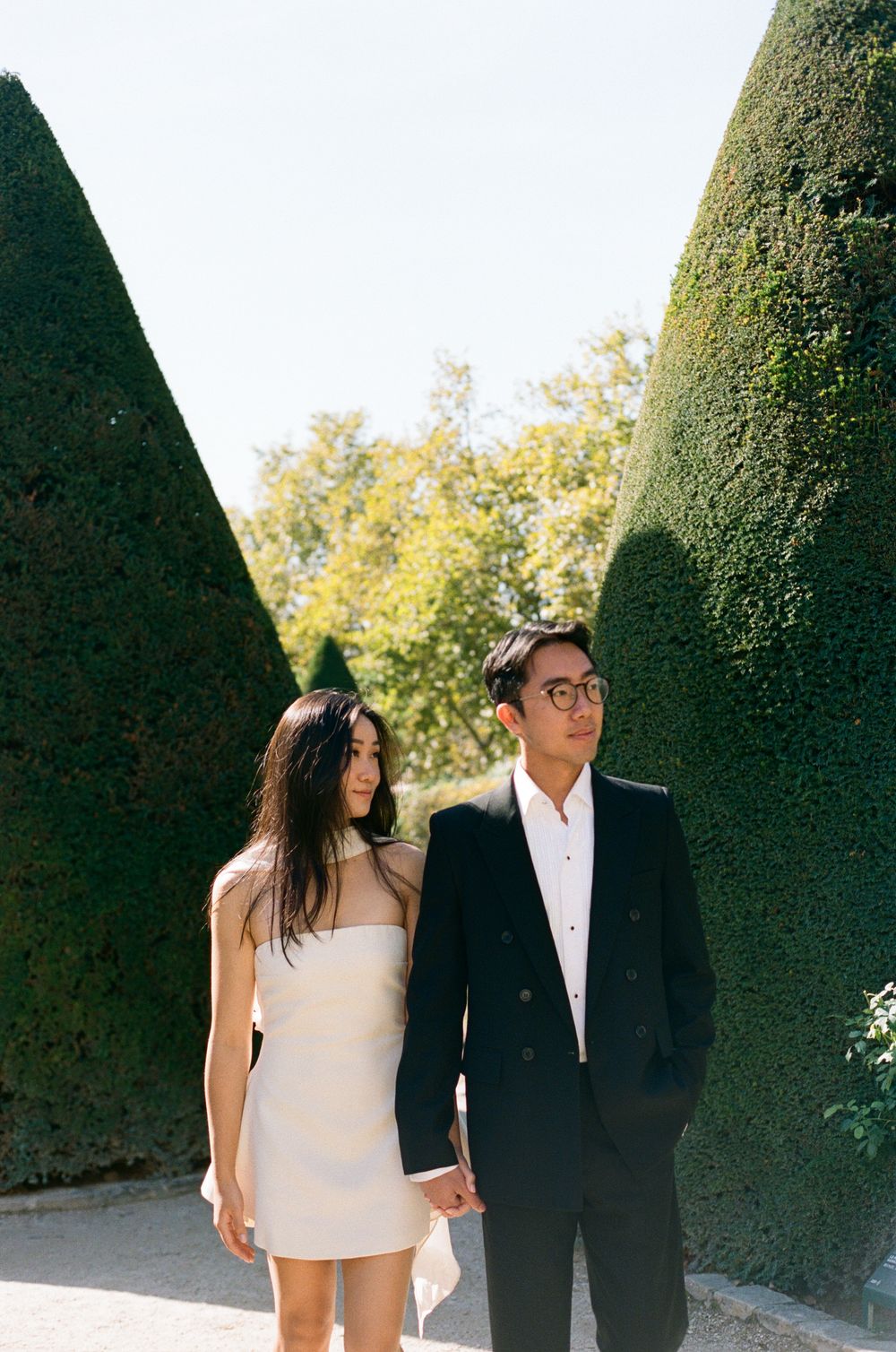 Couple holding hands between tall sculpted hedges in a formal garden, woman in white dress, man in black suit. best paris film photographer