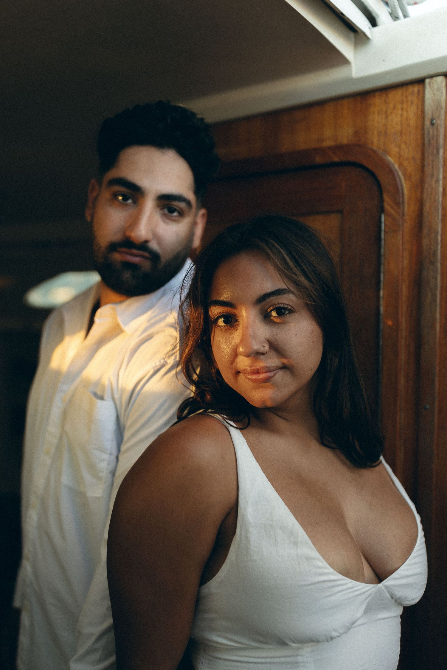 A couple stands together indoors wearing white clothing in warm ambient lighting.