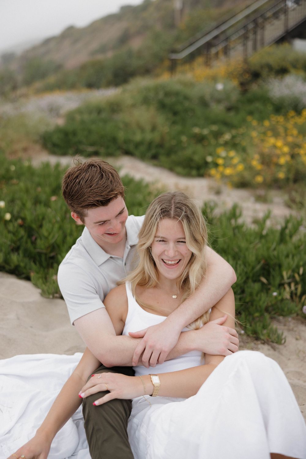 A couple sitting together and laughing on a sandy path surrounded by green coastal vegetation and yellow wildflowers.