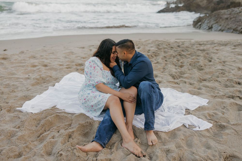 A romantic couple sits intimately together on a white blanket at a scenic beach with crashing waves in the background.