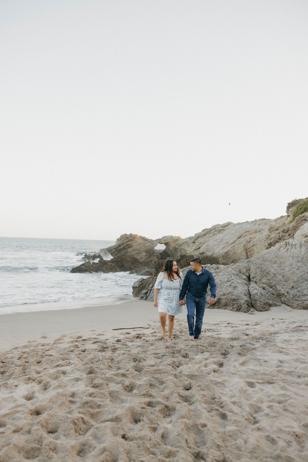 A couple walking together on a scenic beach with rocky cliffs and ocean waves at sunset.