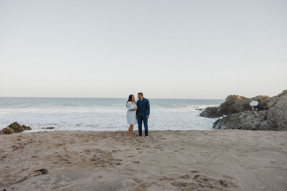 Two figures stand together on a misty beach with rocky outcrops and crashing waves in the background.