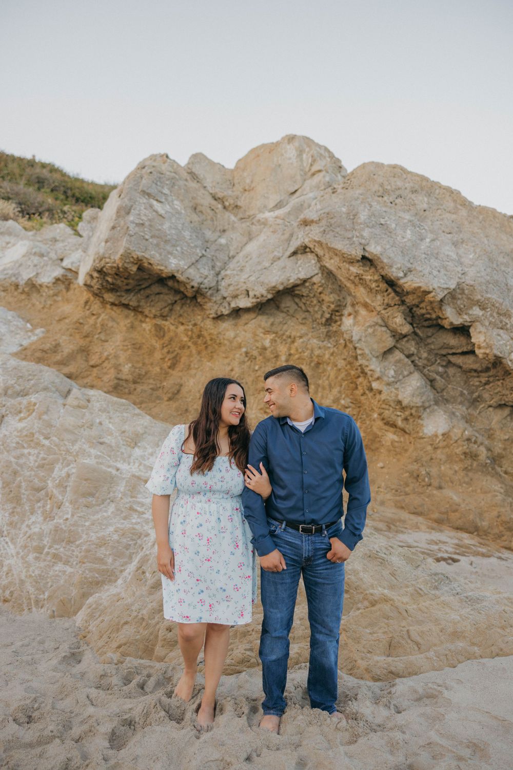 A couple stands embracing on a sandy beach against rugged rock formations and cliffs.