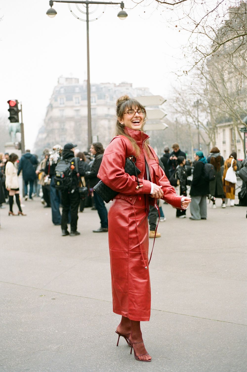 A person in a striking red leather coat and heels poses on a crowded Parisian street during fashion week.