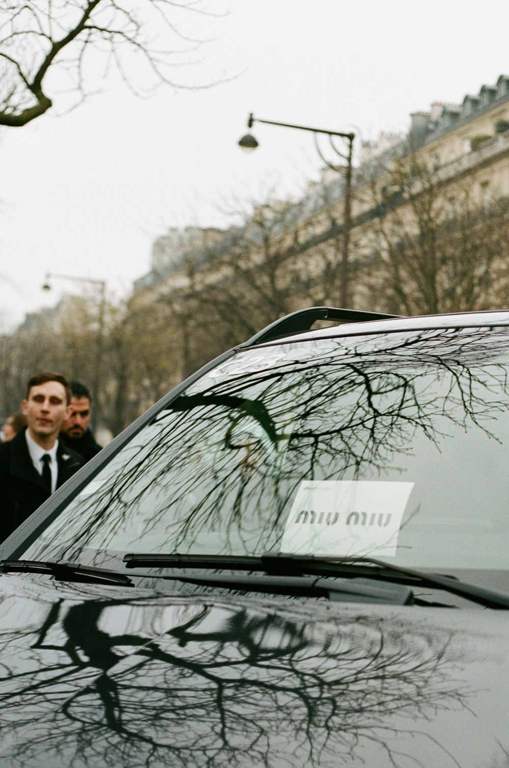 A black luxury car with bare tree branches reflected on its windshield parked on a Parisian street during winter.