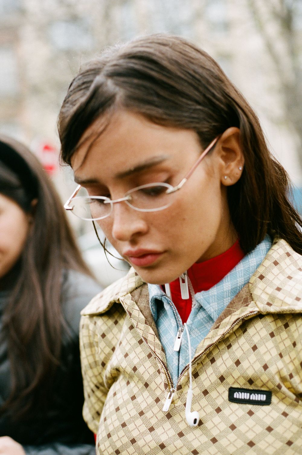 A person wearing frameless glasses and a beige patterned jacket with a red and blue layered collar.