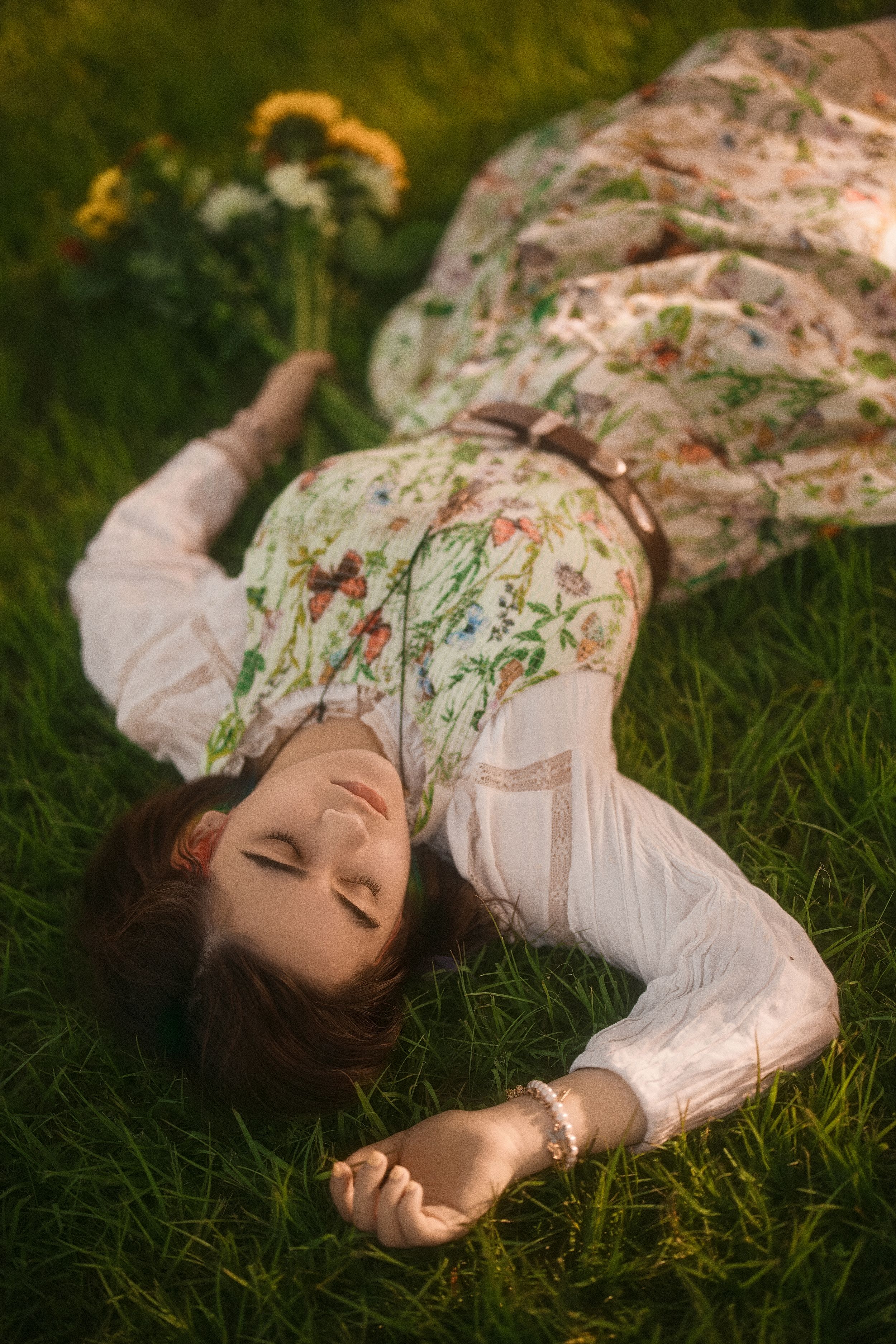 Woman in floral dress lying on green grass with eyes closed, holding wildflowers in warm sunlight.