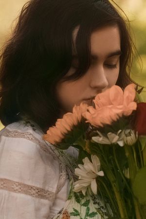 Young woman with dark hair smelling a bouquet of peach and white flowers outdoors.
