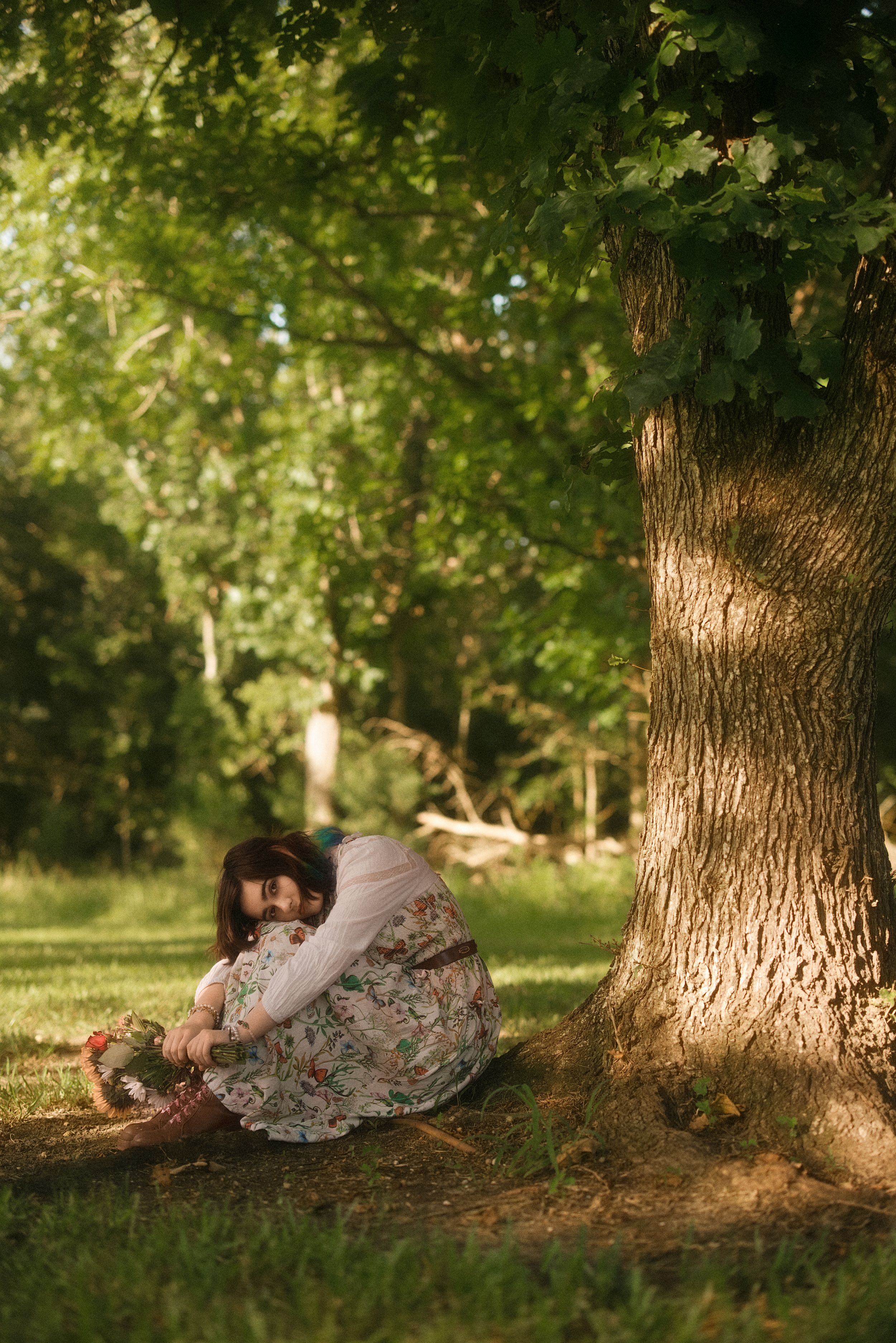 Woman in floral dress sitting under a large oak tree in a sunlit green park, holding a wicker basket.