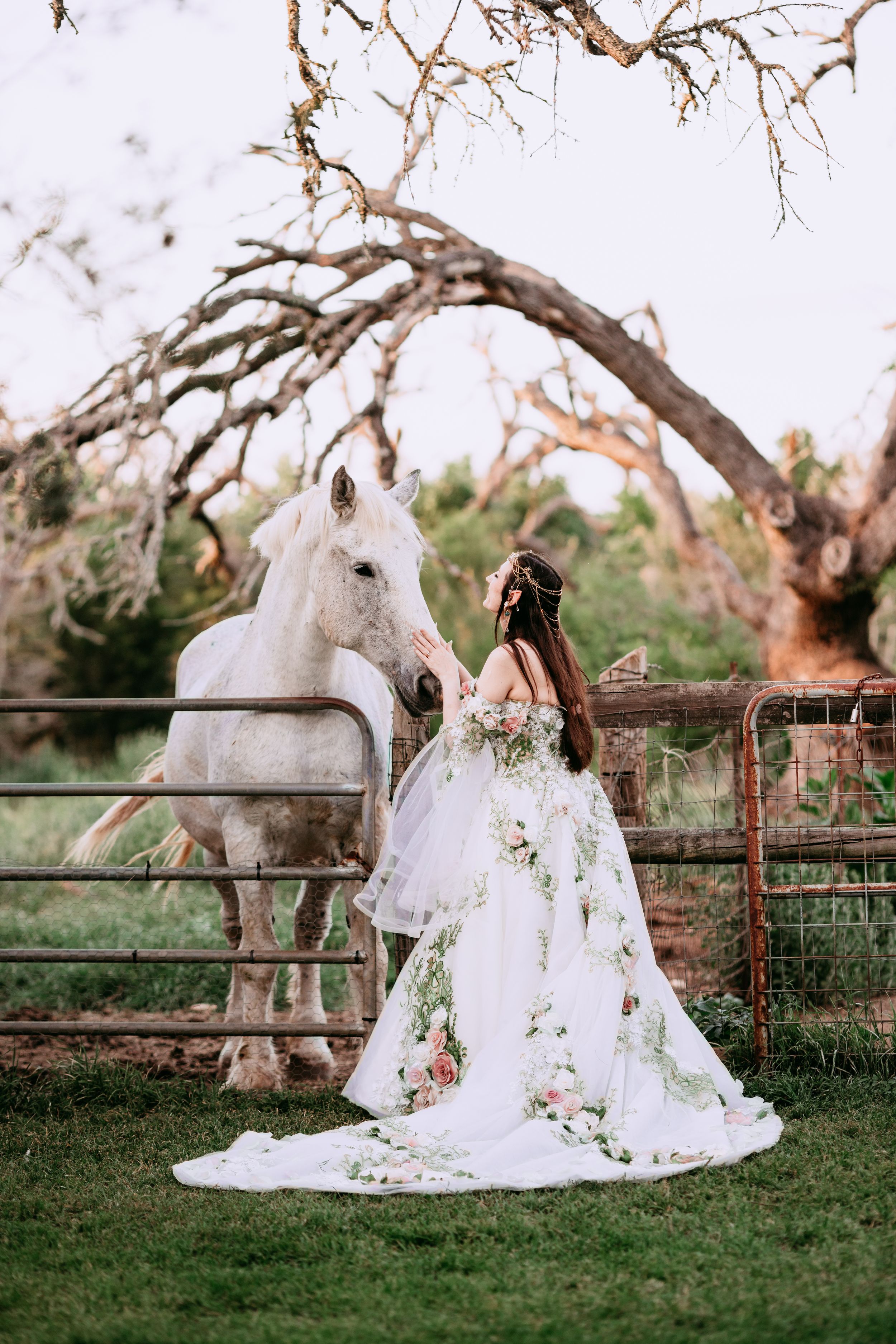 A flowing white wedding dress with floral embellishments is elegantly displayed against a rustic wooden fence at sunset.