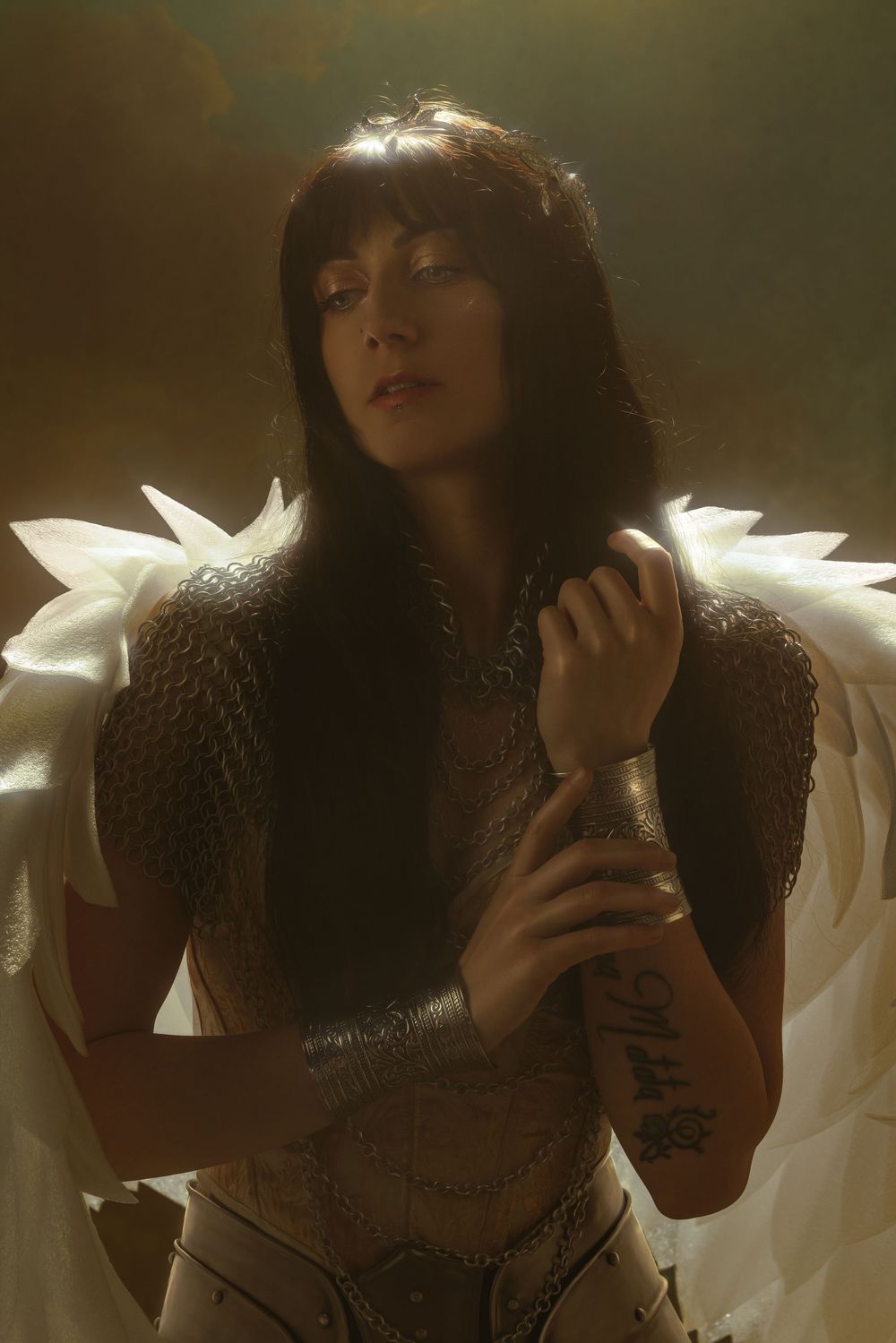 Woman in chainmail armor with white feathered wings and leather bracers poses dramatically in moody golden light.